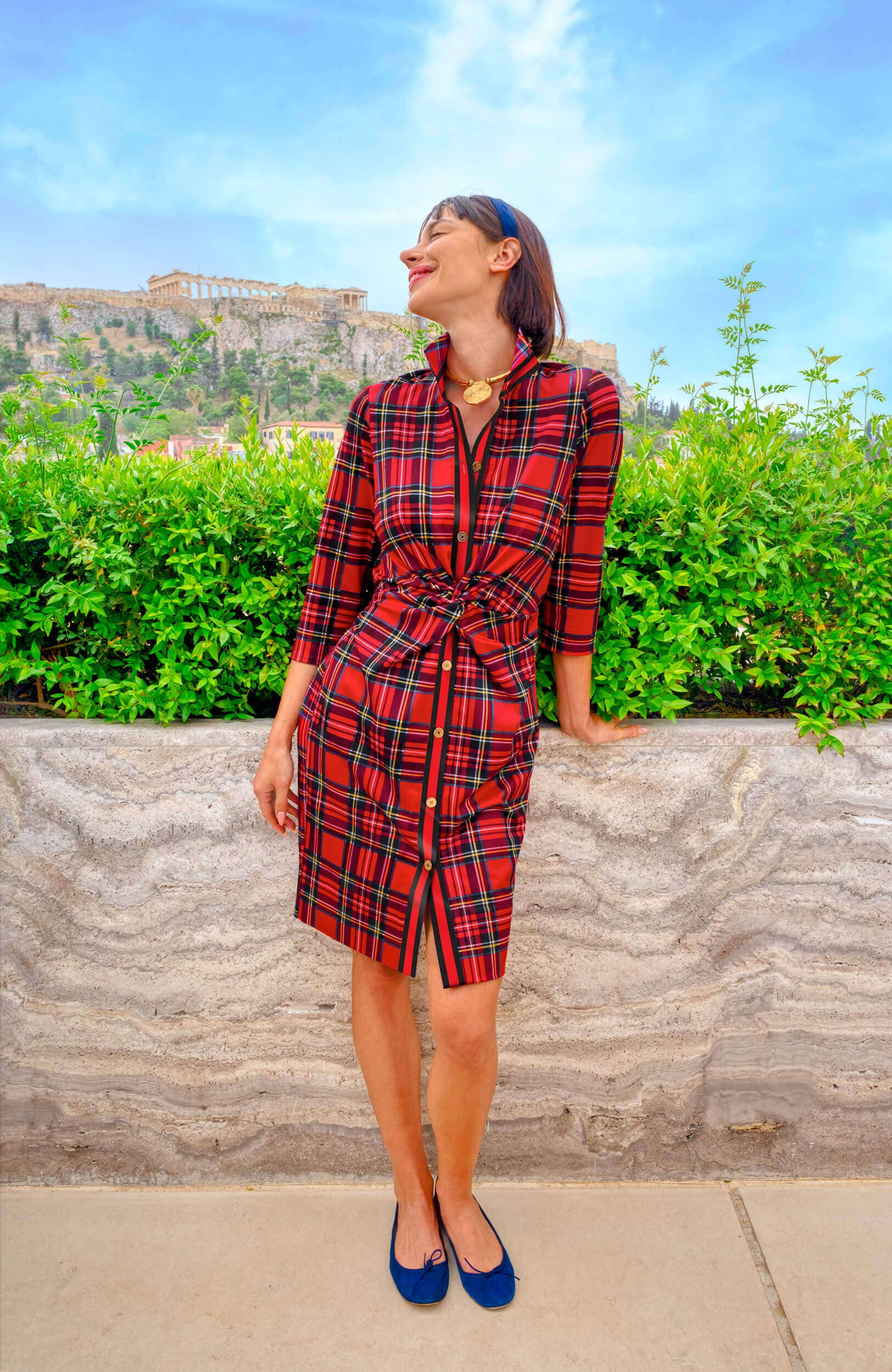 Woman in a red plaid dress standing outdoors with greenery and a building in the background