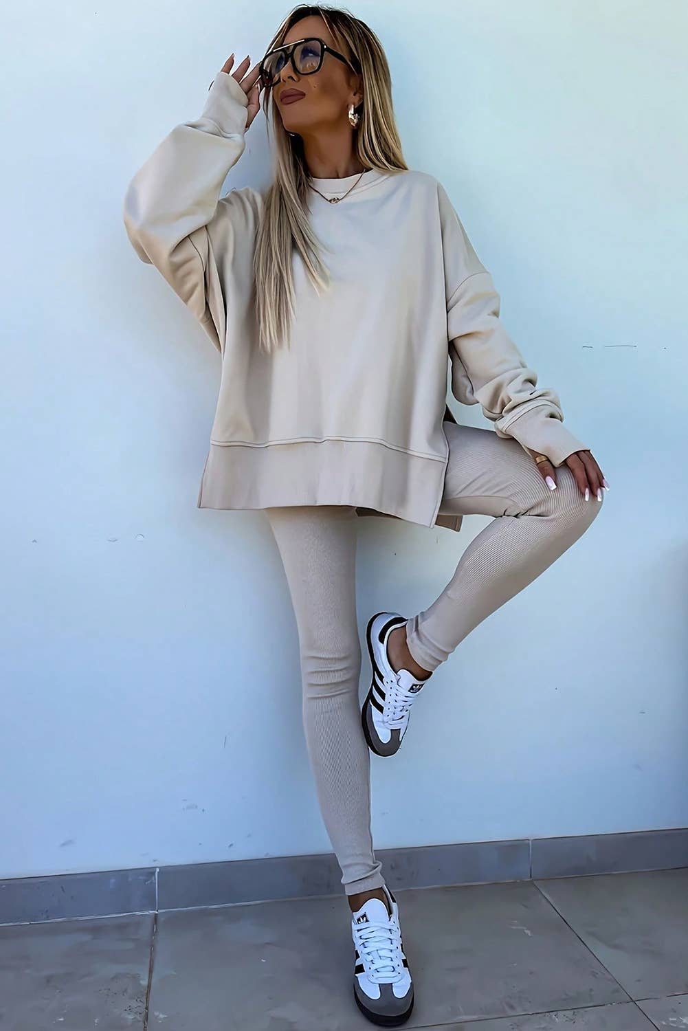 Woman wearing a beige outfit with leggings and sneakers against a light blue wall.